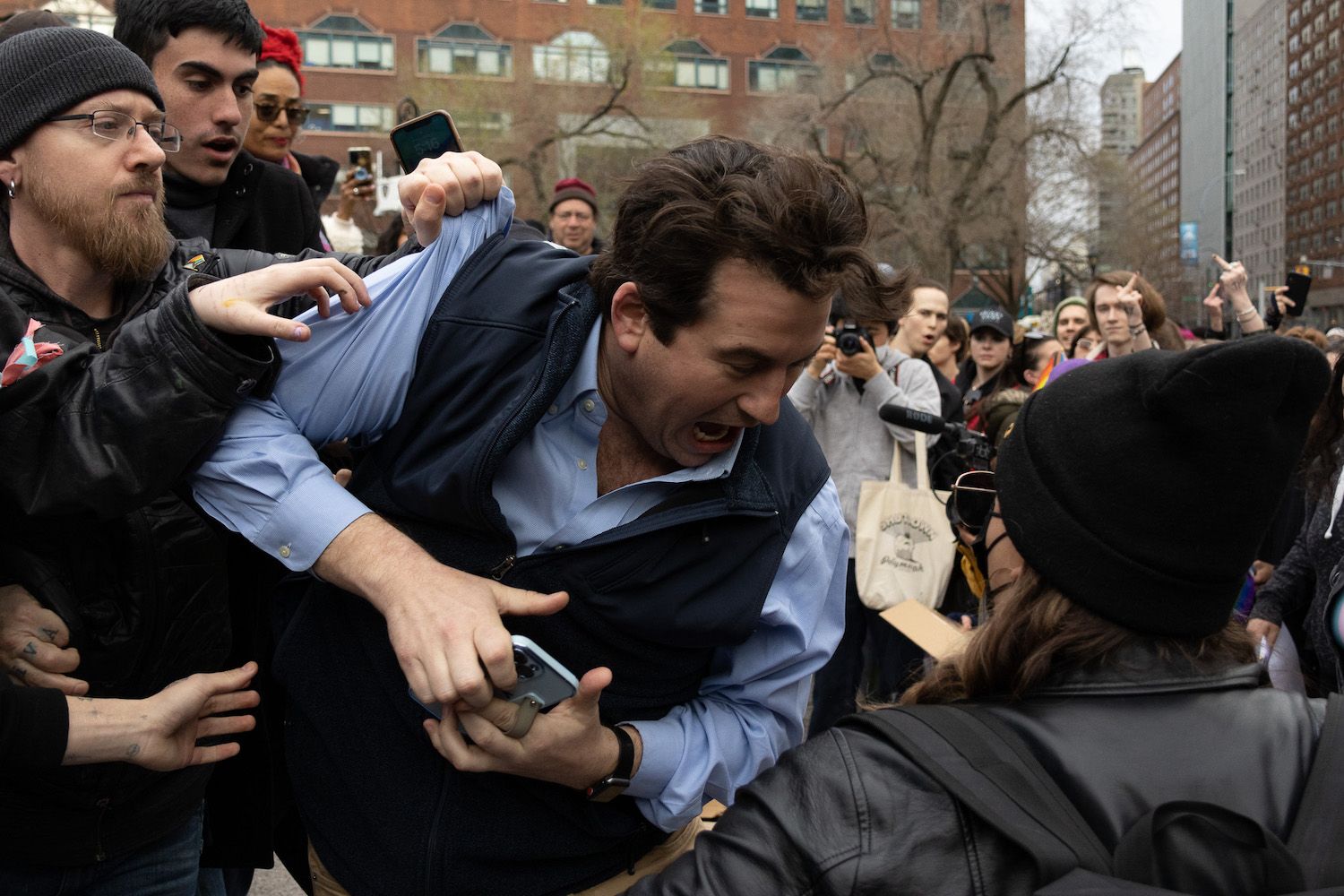 A person wearing a blue shirt and vest and holding a phone makes a distressed face as another man grabs him by the shoulder. Onlookers in the crowd are shocked, calm, or angry.