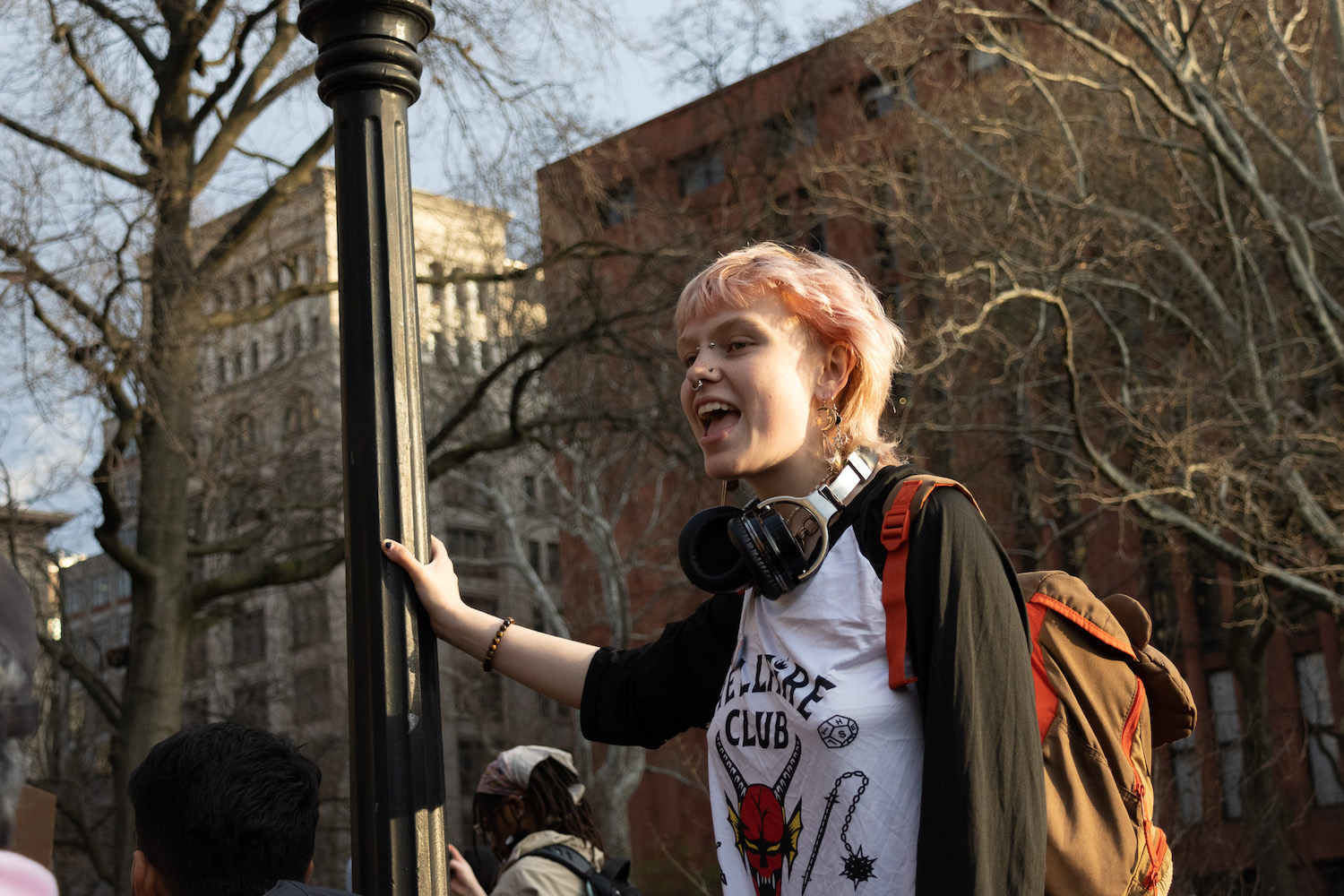 A young person with pink hair and facial piercings stands above a crowd in a park, holding onto a lamppost. Their mouth is open as they chant.