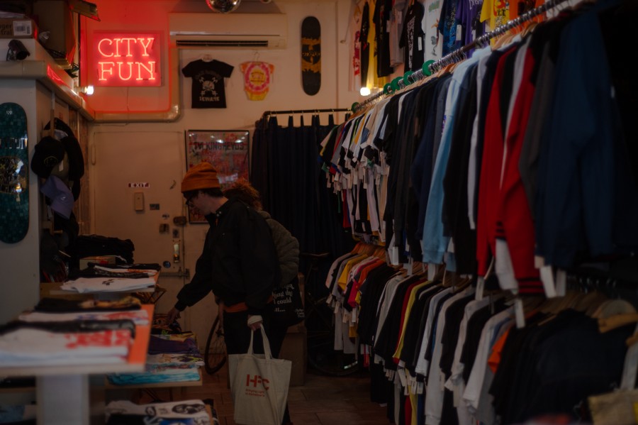 Two+people+browsing+racks+and+tables+of+tee+shirts+in+a+store.+A+red+neon+sign+that+reads+%E2%80%9CCity+Fun%E2%80%9D+hangs+above%2C+and+tee+shirts+and+a+skateboard+hang+from+the+back+wall.