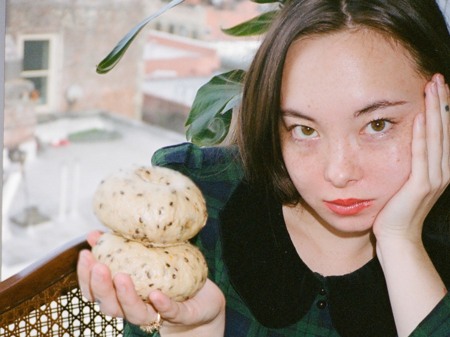 Baker Sakura Smith holds two bagels with her right hand while holding her head with her left hand. She is wearing a green shirt and looking into the camera.