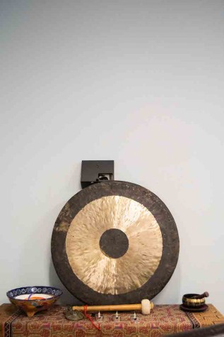 A gong, a mallet and two bowls sit on embroidered fabric covering a table. The table is pushed up against a white wall.