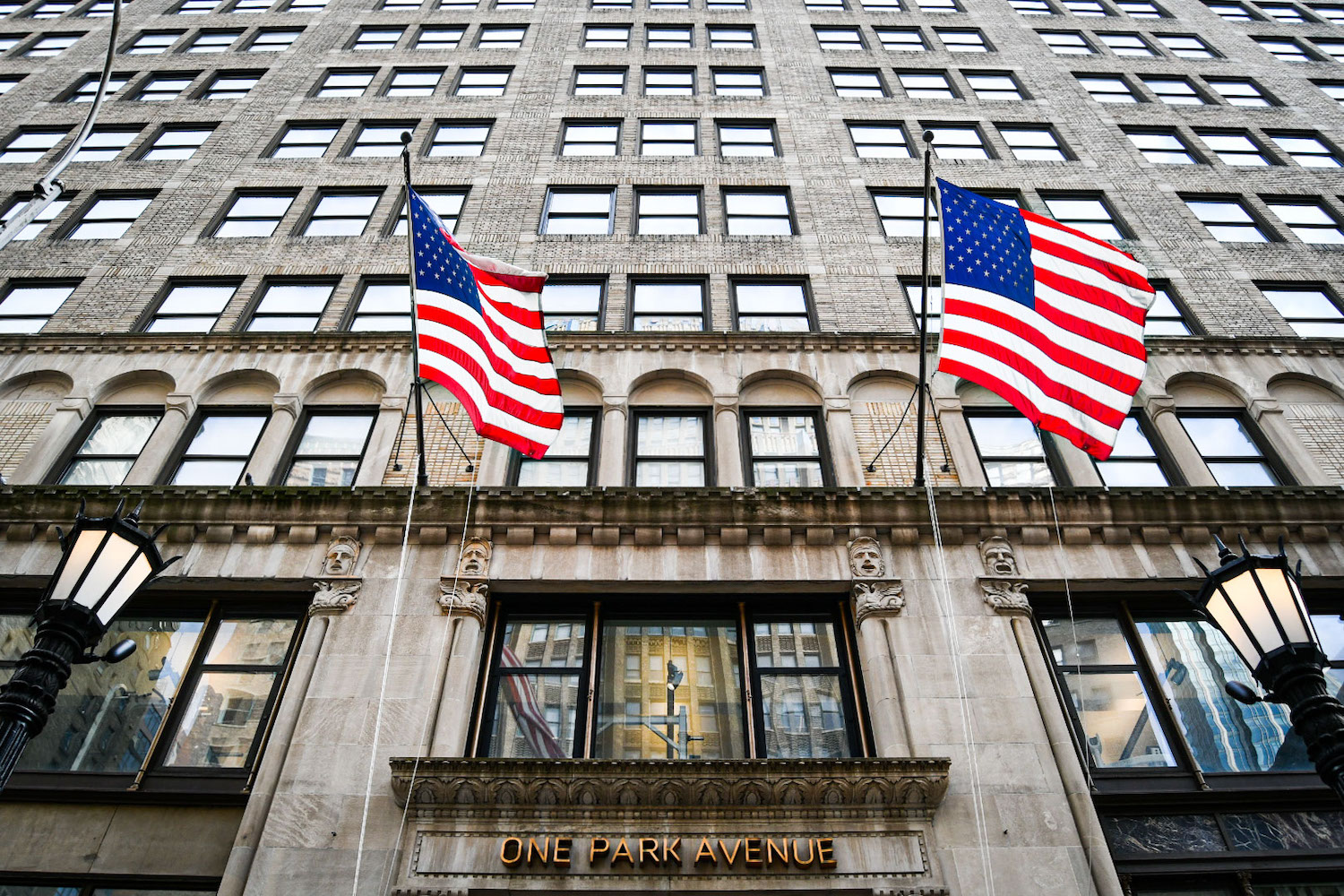 Two American flags hang from the exterior of a building marked as One Park Avenue. The building is partially made of bricks, and has small embedded columns and engraved faces.