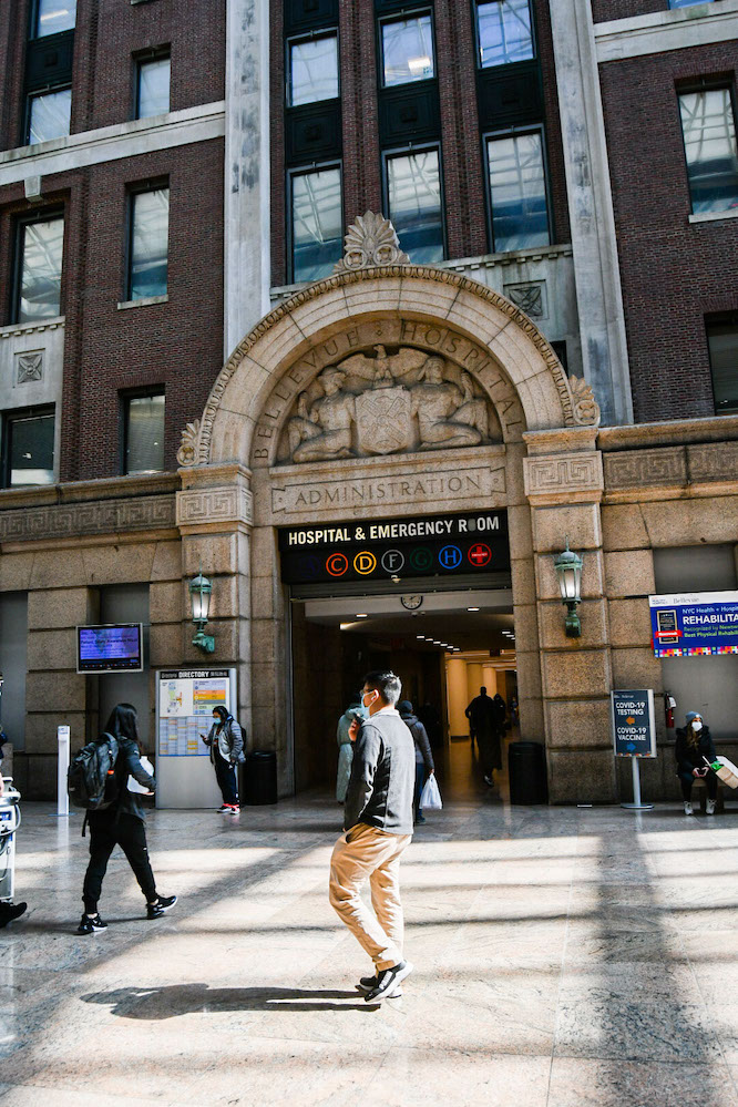 A man in a dark shirt and khaki pants walks on a shiny tile floor outside a hospital building. Two signs hang above the doorway that read “Bellevue Hospital Administration” and “Hospital and Emergency Room.”