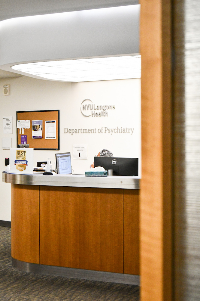 A woman sits behind a computer at the reception desk under a sign that reads “Department of Psychiatry.” A bulletin board is hanging on the wall behind her.