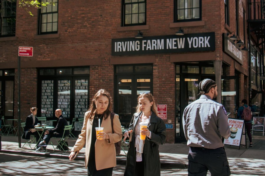 Two women engaged in conversation cross the street as an NYU Campus Safety officer passes by. A sign in the background reads “Irving Farm New York” in white uppercase letters.