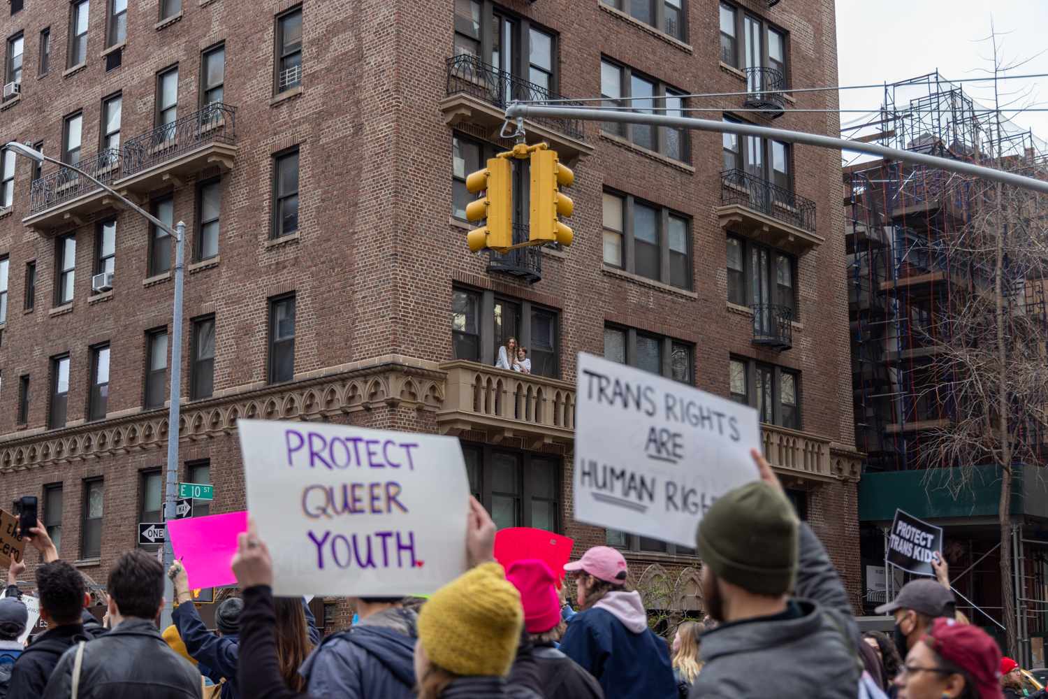 Signs that read “Protect Queer Youth,” and “Trans Rights are Human Rights” held up in front of a brown apartment building with two people looking out on a balcony.