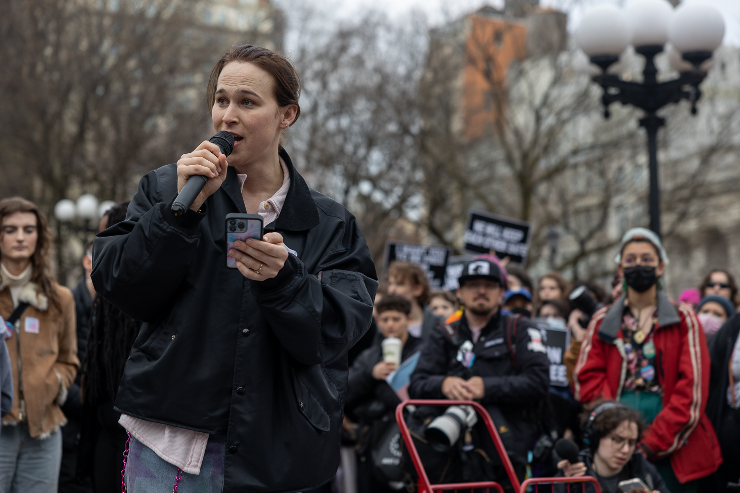 Actress Tommy Dorfman standing and speaking into a microphone with a crowd of people behind her.