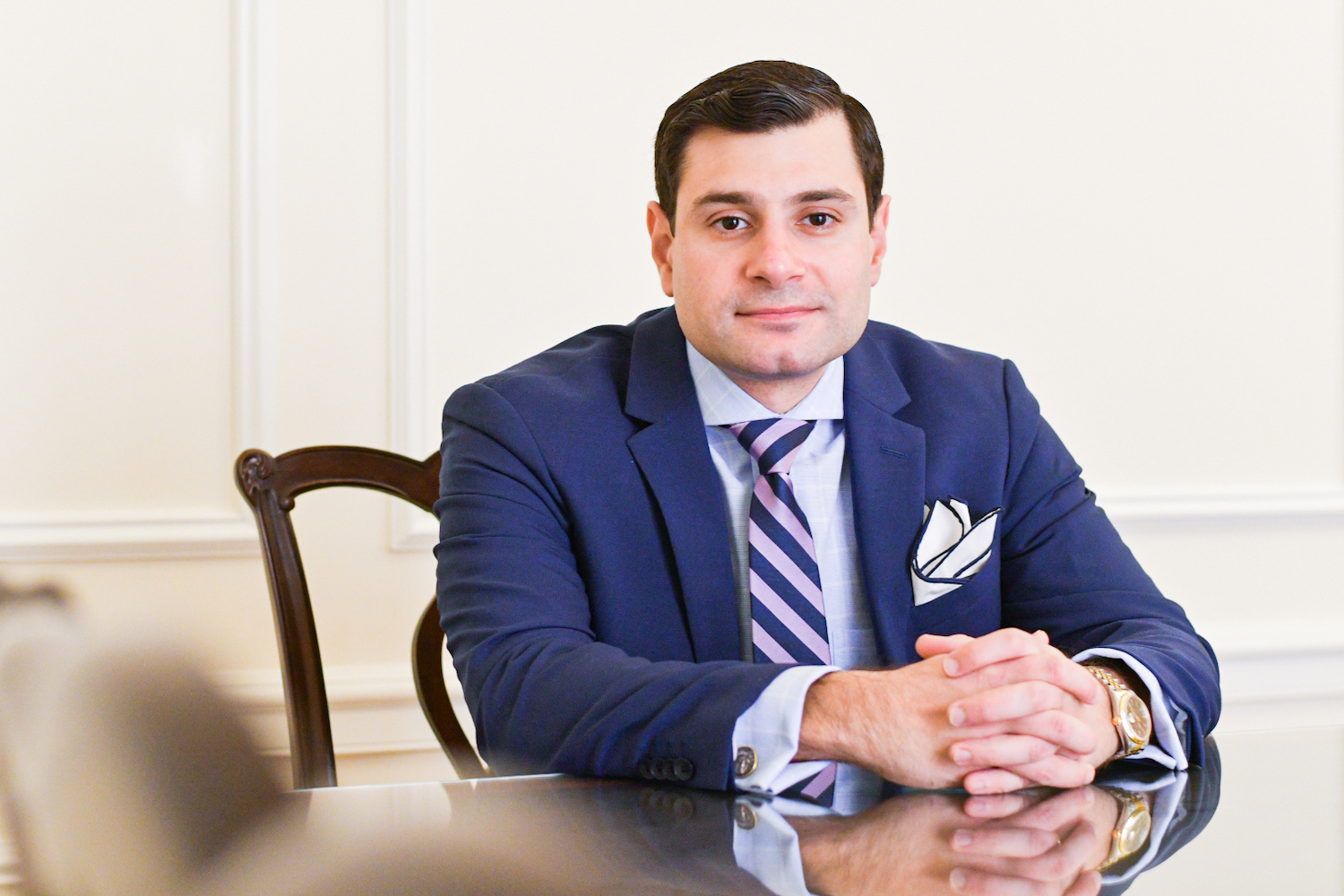 A man with short, brown hair in a dark blue suit and a blue and purple tie sits at a conference table with his hands clasped in front of him. He is looking into the camera.