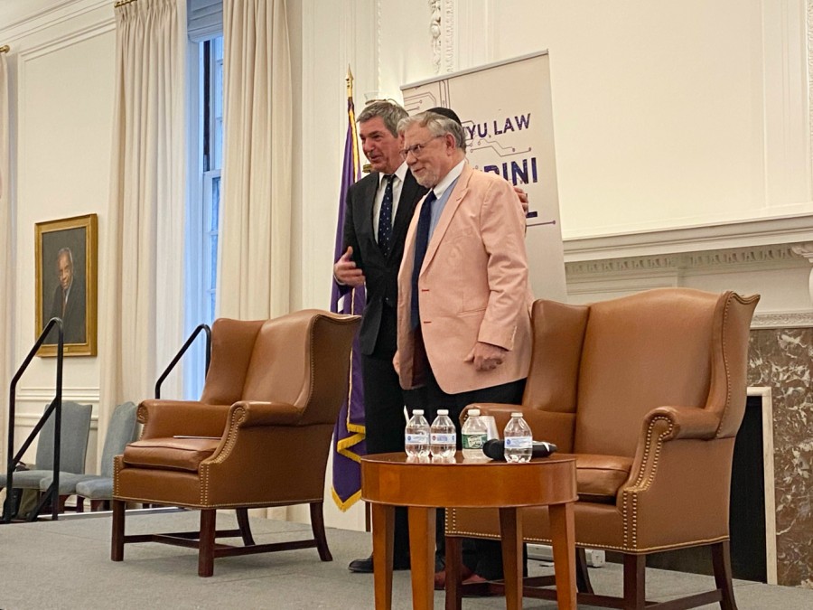 E.U. ambassador Stavros Lambrinidis and N.Y.U. Law professor J.H.H. Weiler standing next to each other on a stage. They are standing next to two large brown armchairs and a small table with water bottles on top.