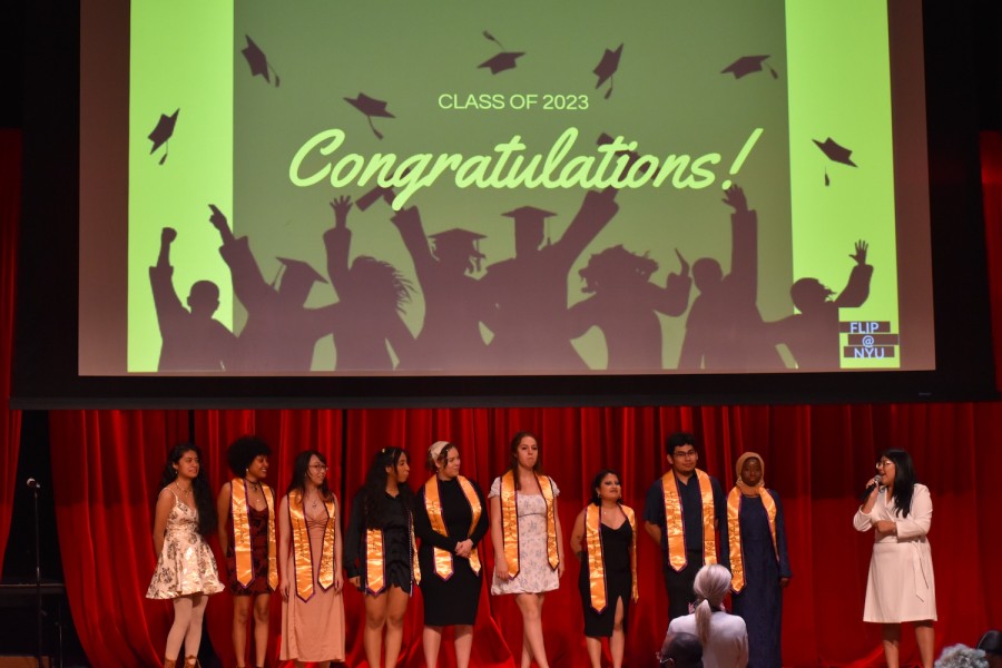 A line of people, most wearing identical golden and purple sashes, stand on a stage in front of red curtains below a large screen. Projected on the screen is an image that reads “CLASS OF 2023” and “CONGRATULATIONS!” with silhouettes of people throwing graduation caps in the air and the phrase “F.L.I.P. @ N.Y.U.” in the corner.