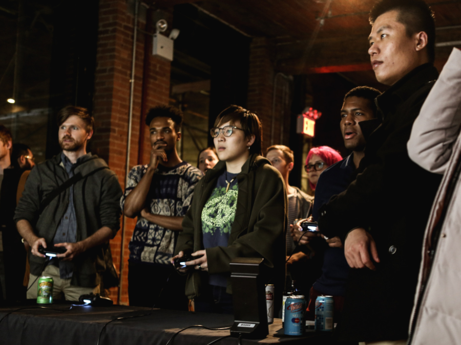 A group of game design students gathers inside a brick building facing towards a screen holding controllers.