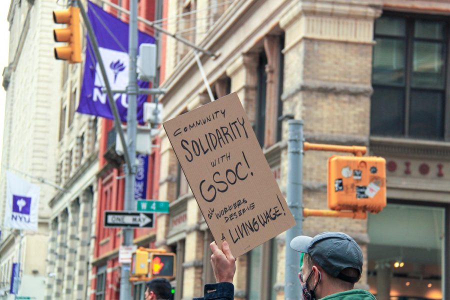 A person holding up a paperboard sign that reads “COMMUNITY SOLIDARITY WITH G.S.O.C.” There is a purple flag of N.Y.U hanging from a building in the background.