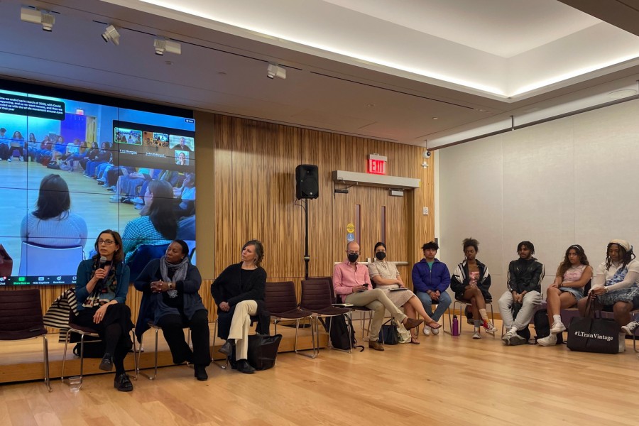 A group of people sitting in a circle in the middle of a conference room with a wooden floor and wall. The display on the wall shows an online conference call.