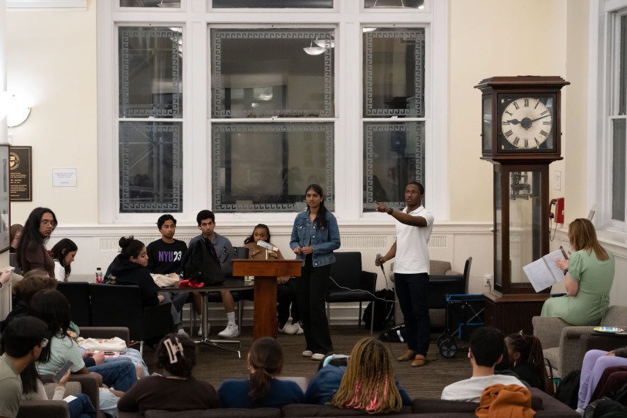 A group of people sitting in a room with a clock on the side. Two speakers stand in front of the crowd with one of them holding a microphone while pointing to the crowd.