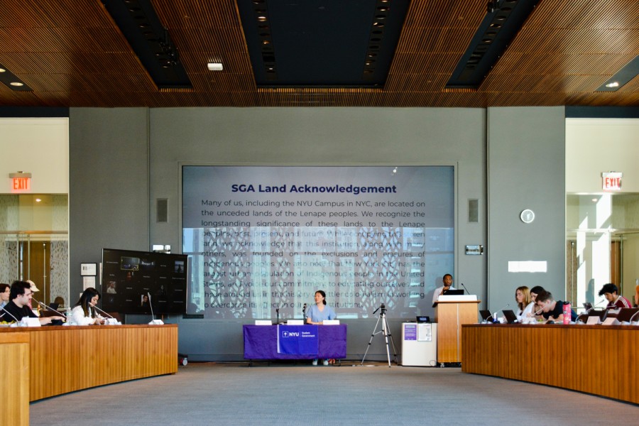 A group of people sits around a purple table in the middle of a room with a podium at the center. A person is speaking behind the podium. Behind the purple table is a screen displaying the text “S.G.A. Land Acknowledgment.”