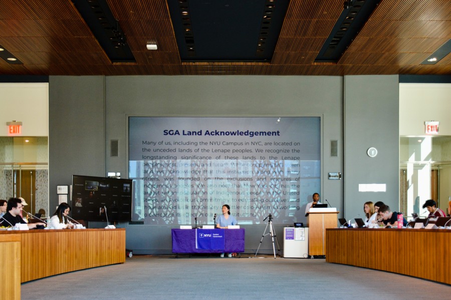 A+group+of+people+sits+around+a+purple+table+in+the+middle+of+a+room+with+a+podium+at+the+center.+A+person+is+speaking+behind+the+podium.+Behind+the+purple+table+is+a+screen+displaying+the+text+%E2%80%9CS.G.A.+Land+Acknowledgment.%E2%80%9D