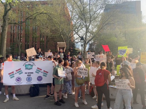 A crowd gathered at Washington Square Park. People are holding various colored signs that say “FUCK YOU, PAY US!” and “MAKE SOCIAL WORK ACCESSIBLE!” among others. Closest to the camera are two people who hold a banner that reads “2023 P4P NYC RALLY – PAY INTERNS NOW!”