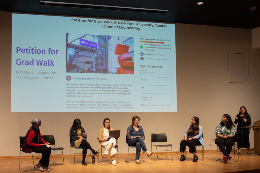 Six people sit on a stage with a projector screen behind them displaying an online petition for the graduation walk for N.Y.U.’s Tandon School of Engineering. There is a speaker holding a microphone on the right.