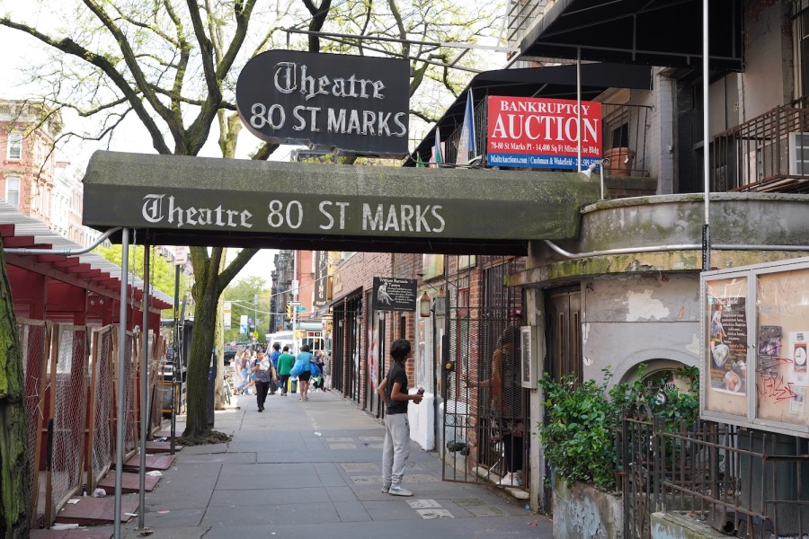 The entrance to Theatre Eighty Saint Marks with a person standing outside. An old-fashioned green awning hangs above the entrance and a bright red sign hanging on the building reads, “Bankruptcy” and “Auction.”