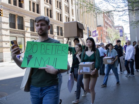 A line of people walk on the street outside N.Y.U.’s Bobst Library. The person in the front of the line is holding a green sign that reads “DO BETTER N.Y.U.”