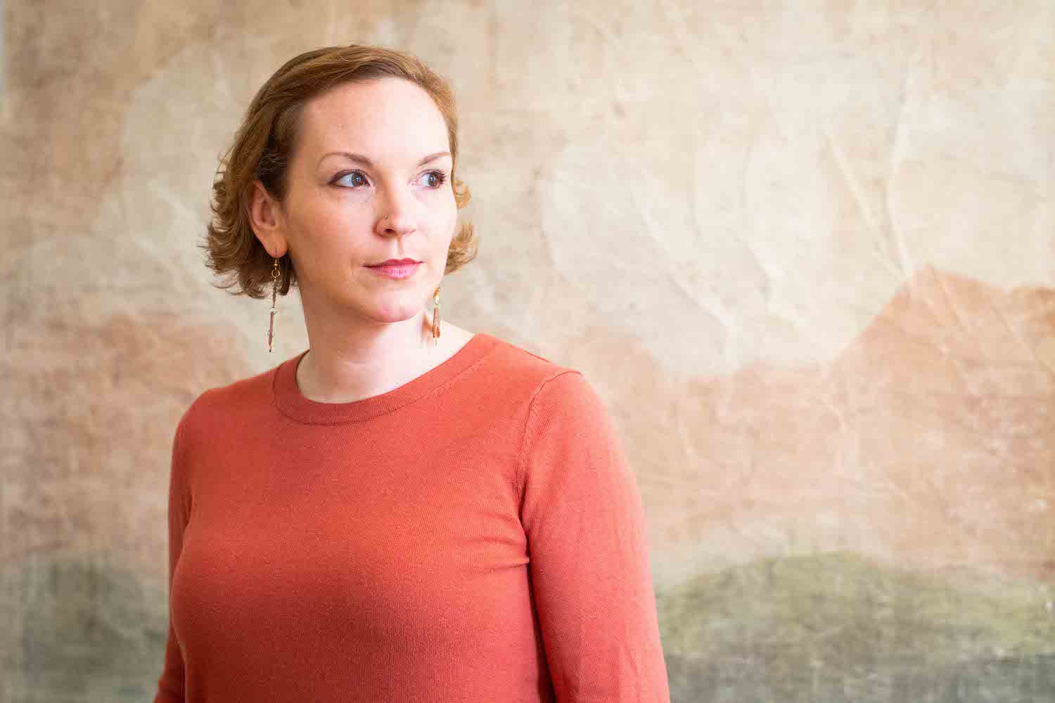 A woman in an orange top, with earrings, short hair and a nose piercing, looking to the right. She is standing in front of a green and beige backdrop.