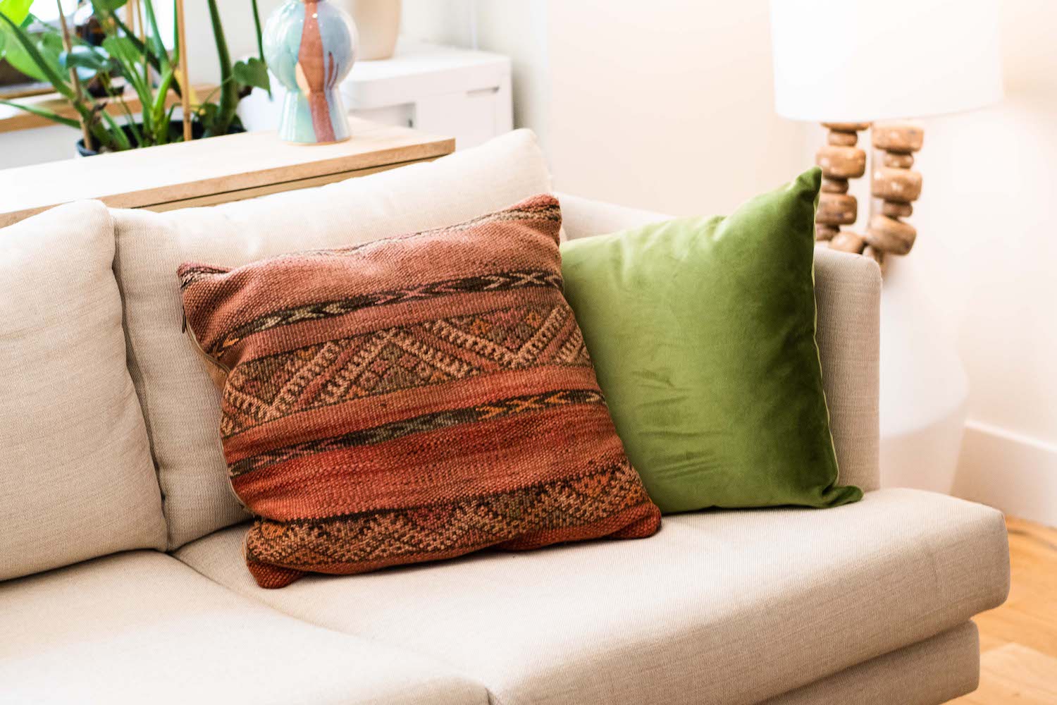 Two pillows, one orange and patterned and the other green, sitting in the corner of a beige couch in a brightly lit room.