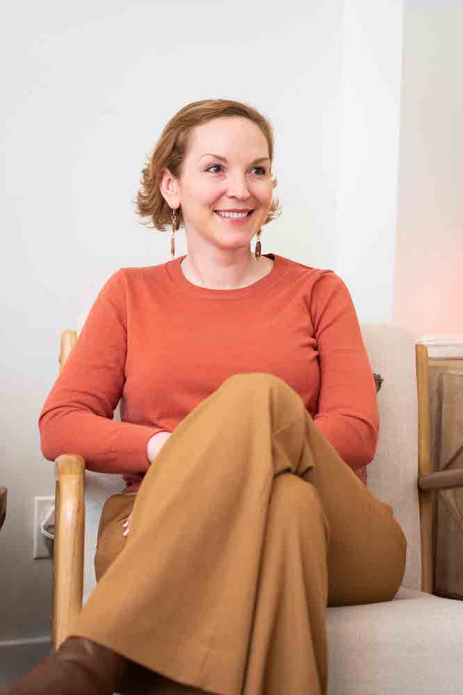 A woman in an orange sweater and light brown pants sits in a chair while smiling and looking to the right. Her legs are crossed.