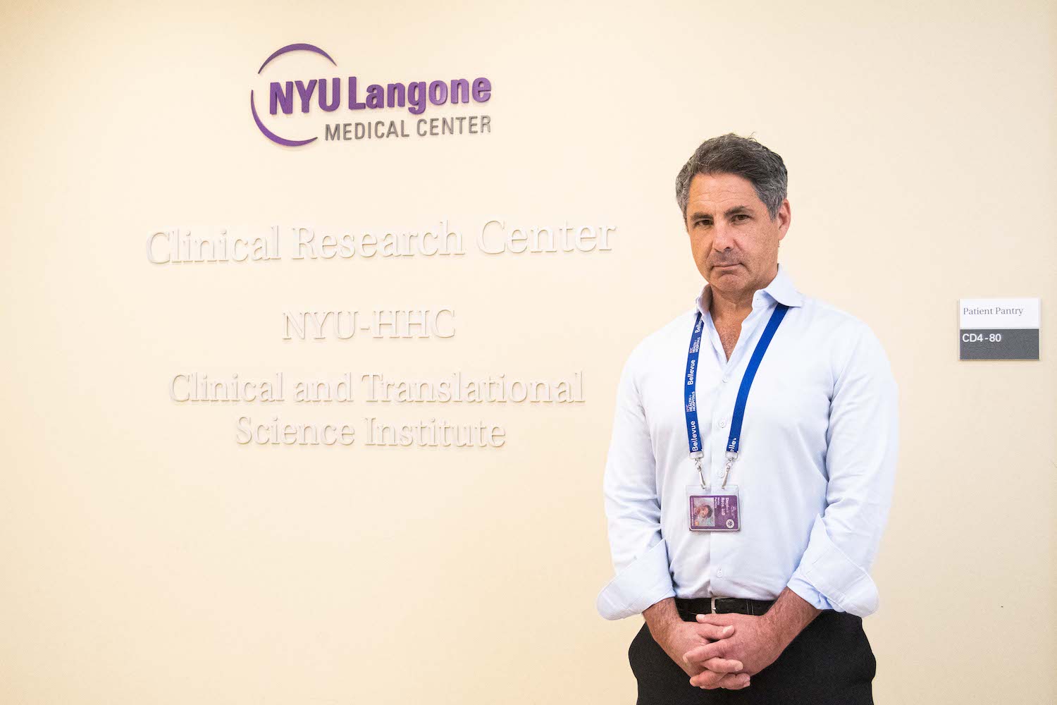 A man wearing a light blue dress shirt and a lanyard around his neck holding an N.Y.U. I.D. stands with his hands clasped in front of him. He is looking into the camera. Behind him is a sign that reads, “N.Y.U. Langone Medical Center,” “N.Y.U. H.H.C.,” “Clinical and Translational Science Institute” and “Clinical Research Center.”