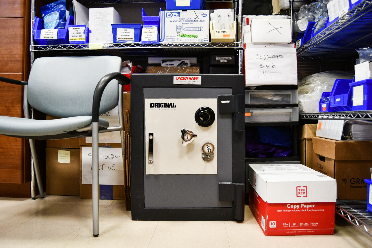A safe placed next to a chair and a paper box on the ground with shelves of miscellaneous items in the background.