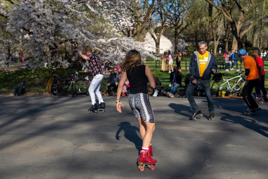 A group of people roller skate in Central Park, the person in the front wears a black top, rolled up pants, and red skates. The skaters in the back wear jeans, t-shirts and black skates.