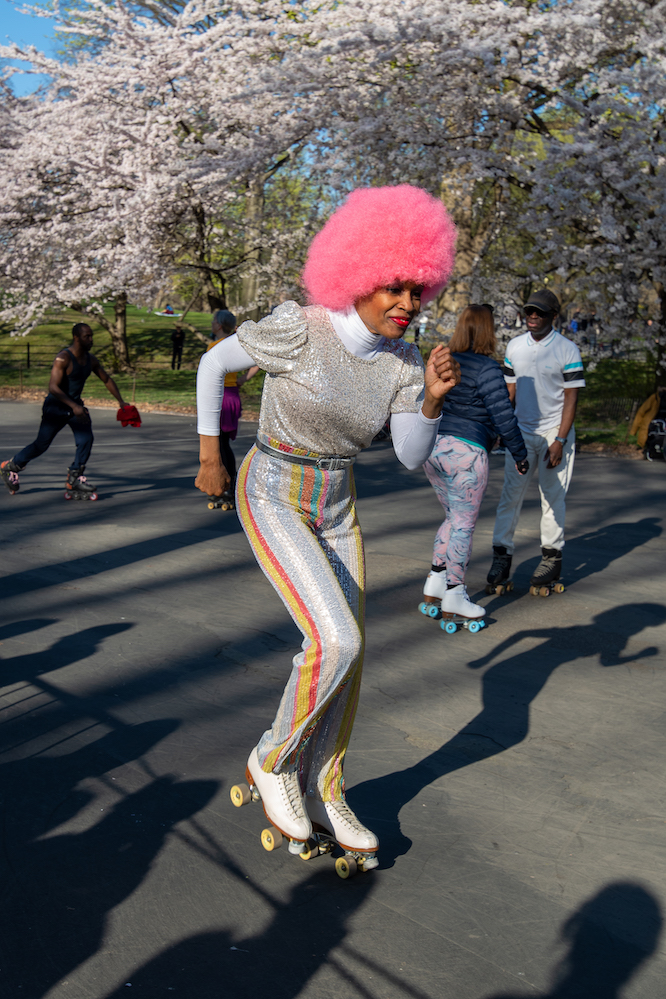 A person wearing a silver, sequined outfit and a bright pink curly wig roller skates while striking a pose in Central Park. Other skaters are in the background on a sunny day.