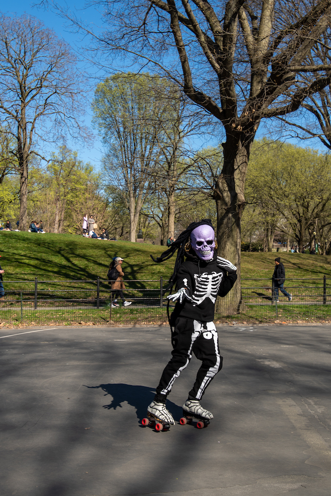 A person wearing a purple skeleton mask, long-haired wig and a skeleton outfit roller skates while flipping their hair and posing in Central Park on a sunny day.
