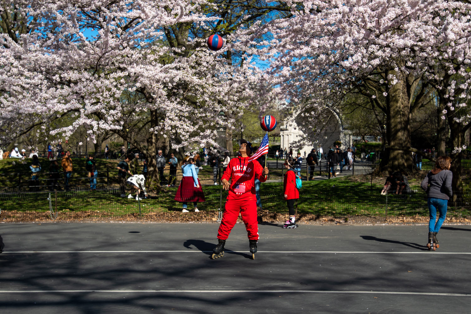 A person wearing an all-red sweatsuit with "Ricky Superstar" printed on the front spins a basketball on top of an American flag while throwing another ball in the air. The person is doing this while wearing inline skates.