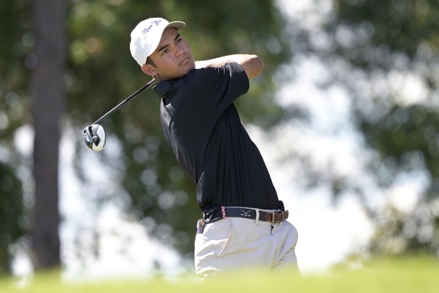 A golf player swings a club. He is wearing a black golf shirt, beige pants and a white cap.