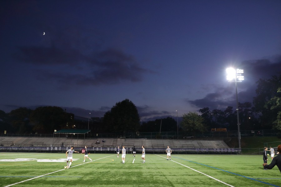 Several soccer players play on a field at night. The lights around the field are on.
