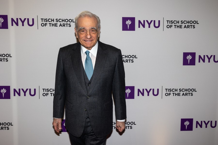 Director Martin Scorsese wearing a suit and a blue tie, standing in front of a backdrop with the N.Y.U. logo and text “TISCH SCHOOL OF THE ARTS.”