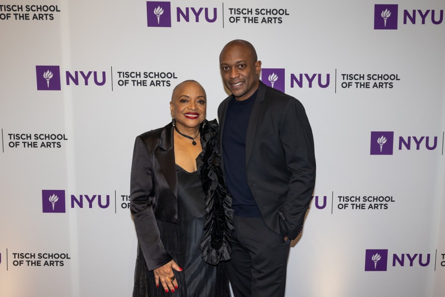 Deborah Willis wearing a black dress and Hank Willis Thomas wearing a black suit, both standing in front of a backdrop with the N.Y.U. logo and text “TISCH SCHOOL OF THE ARTS.”