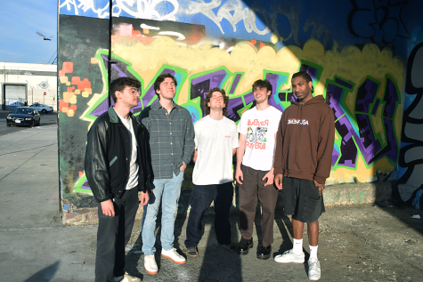 A group of five people standing in a line in front of a graffiti wall, smiling. The sun shines brightly onto their faces.