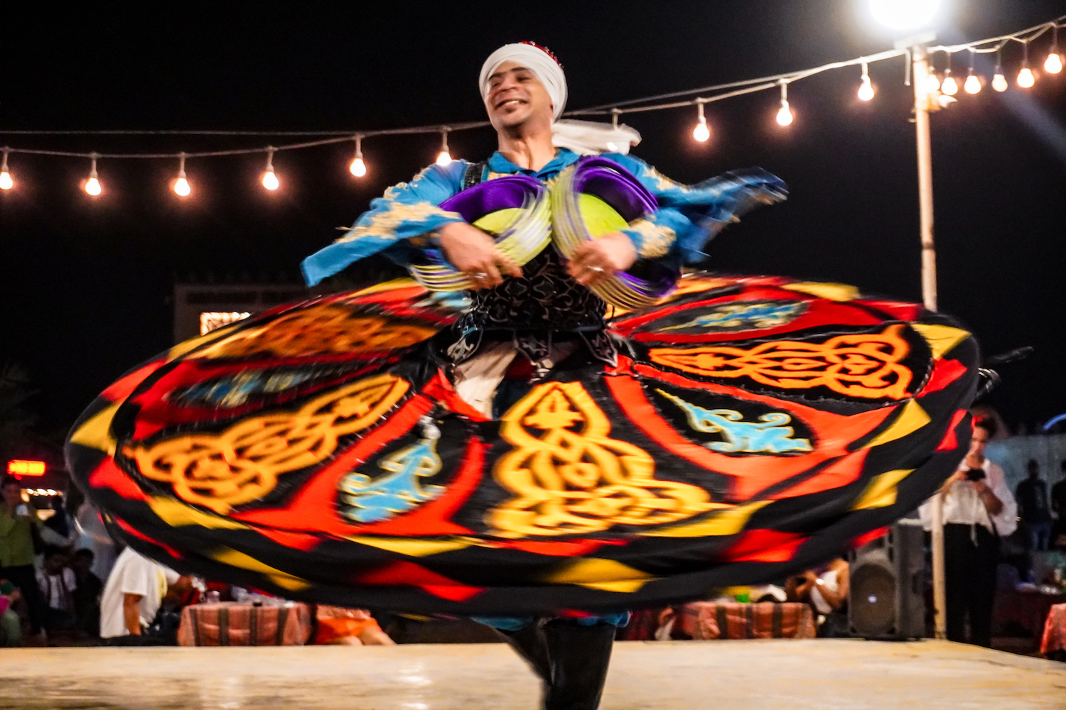 A tanoura dancer in red, yellow, orange, blue and black outfit spins and smiles.