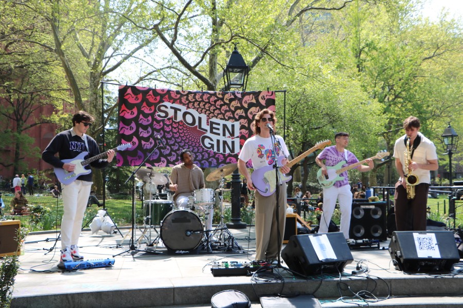 A band performing on a stage with amps in Washington Square Park, with pastel colored guitars, a saxophone and a drum set. Two of the band members wear N.Y.U. merch, and the lead singer is wearing sunglasses. A brightly colored banner with a spiral design hangs behind them, and reads “Stolen Gin,” the name of the band.