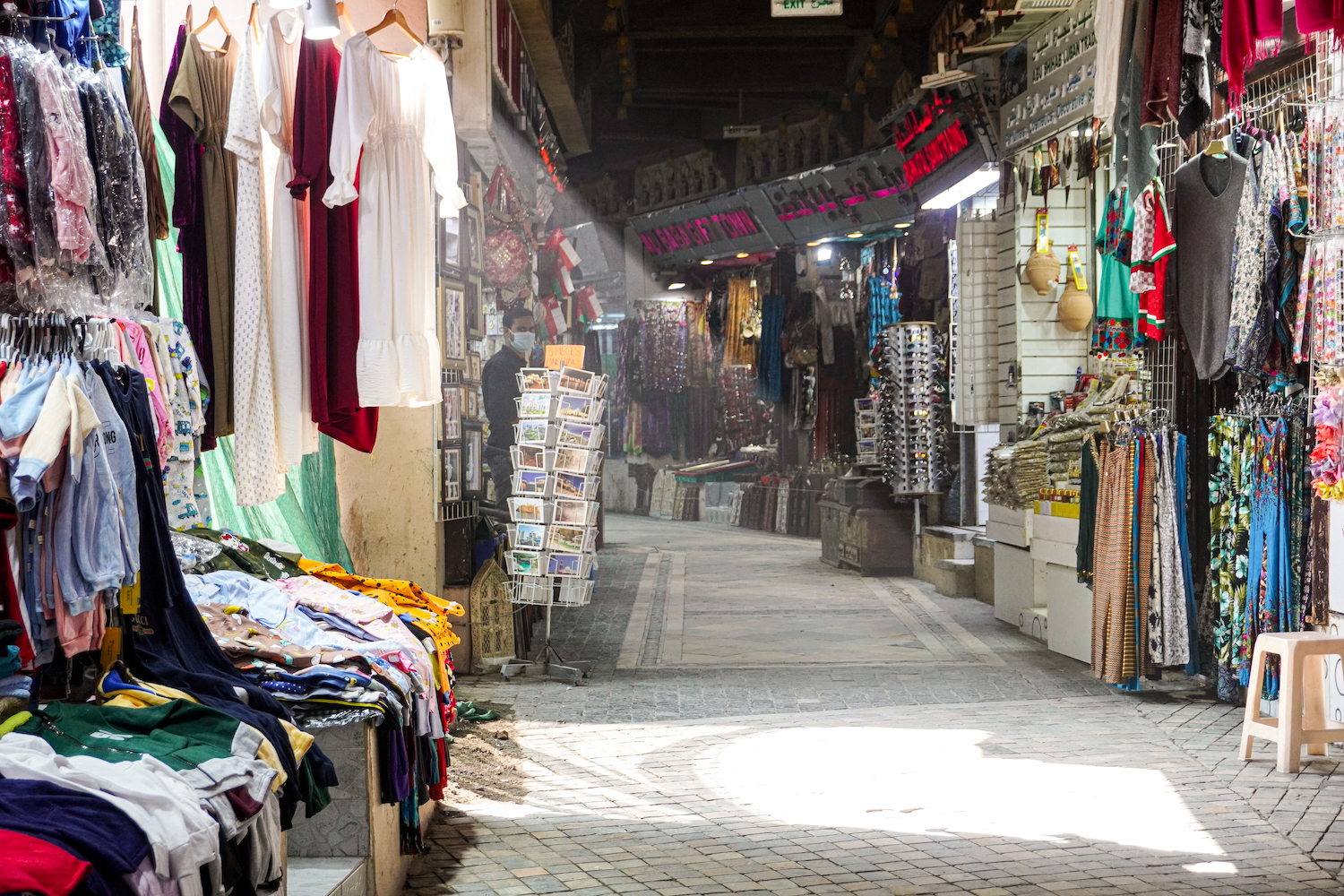 Sunlight pouring into the Mutrah Souq. Many clothes are lined up in stalls along the outdoor market.