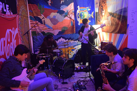 A group of five people with instruments, rehearsing inside a small dimly lit room with bright tapestries covering the walls. Two sit on a dark blue couch, and the others are scattered around the room, seated and standing.
