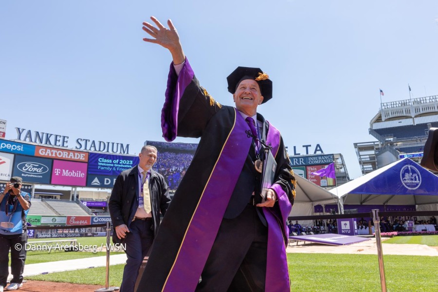 N.Y.U. President Andrew Hamilton standing in a packed Yankee Stadium. He smiles and waves while wearing a black-and-purple robe with gold embellishments.