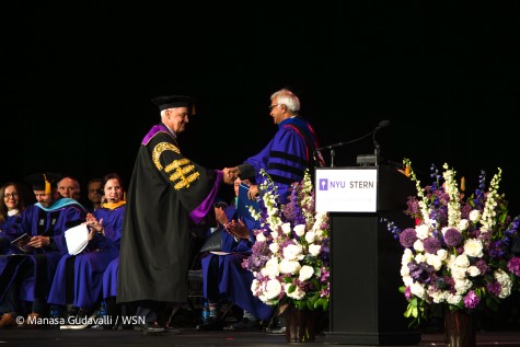 N.Y.U President Andrew Hamilton — wearing a black robe with purple lining and gold embellishments — and Stern dean Raghu Sundaram — wearing a black-and-purple robe with a thin, red lining over his shoulders — shake hands. They are on a stage which has an empty podium with a microphone on top, with the N.Y.U torch and text “N.Y.U Stern Undergraduate College” on it. Flowers surround the podium, and others in various purple robes sit to the left of the handshake, applauding.