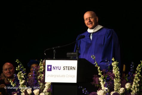 Goldman Sachs C.E.O David Solomon — wearing a purple robe with a white collar — speaks behind a podium with the N.Y.U torch and text “N.Y.U Stern Undergraduate College” on it. Flowers surround him, and one sitting person’s head can be seen in the lower left.