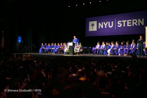 A wide view of the N.Y.U Stern graduation ceremony. Goldman Sachs C.E.O David Solomon — wearing a purple robe with a white collar — speaks behind a podium. Flowers surround him, and people in purple robes sit on stage. There is a full audience watching the stage, most people wearing graduation caps and robes.