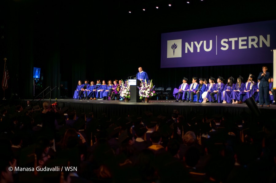 A wide view of the N.Y.U Stern graduation ceremony. Goldman Sachs C.E.O. David Solomon — wearing a purple robe with a white collar — speaks behind a podium. Flowers surround him, and people in purple robes sit on stage. There is a full audience watching the stage, most people wearing graduation caps and robes.