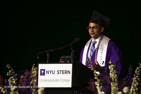 Stern graduate Samyak Sawale, the student speaker, wears a purple robe with a white sash over his shoulders overtop a white button-up shirt and purple tie, and a black graduation cap. He speaks behind a podium with the N.Y.U torch and text “N.Y.U Stern Undergraduate College” on it. Flowers surround him, and one sitting person’s head can be seen in the lower left.