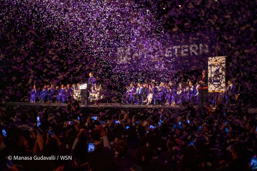 A view of the N.Y.U Stern graduation ceremony. White text reading “STERN” can be seen on the far back wall. A crowded audience, several with phones out recording, cheer, as purple confetti showers them. People in purple robes sit on stage, while one person stands behind a podium.