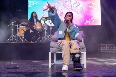 Pavari wears a sky blue sweater covered in white clouds over a yellow top, beige pants and a bedazzled medical boot. She sits on a stack of three gray pillows, and points out towards the crowd with one hand and holds a microphone in the other. Behind her is a screen displaying her name in wavy font over a blue-and-pink background. A man with a drum set sits on a raised platform.